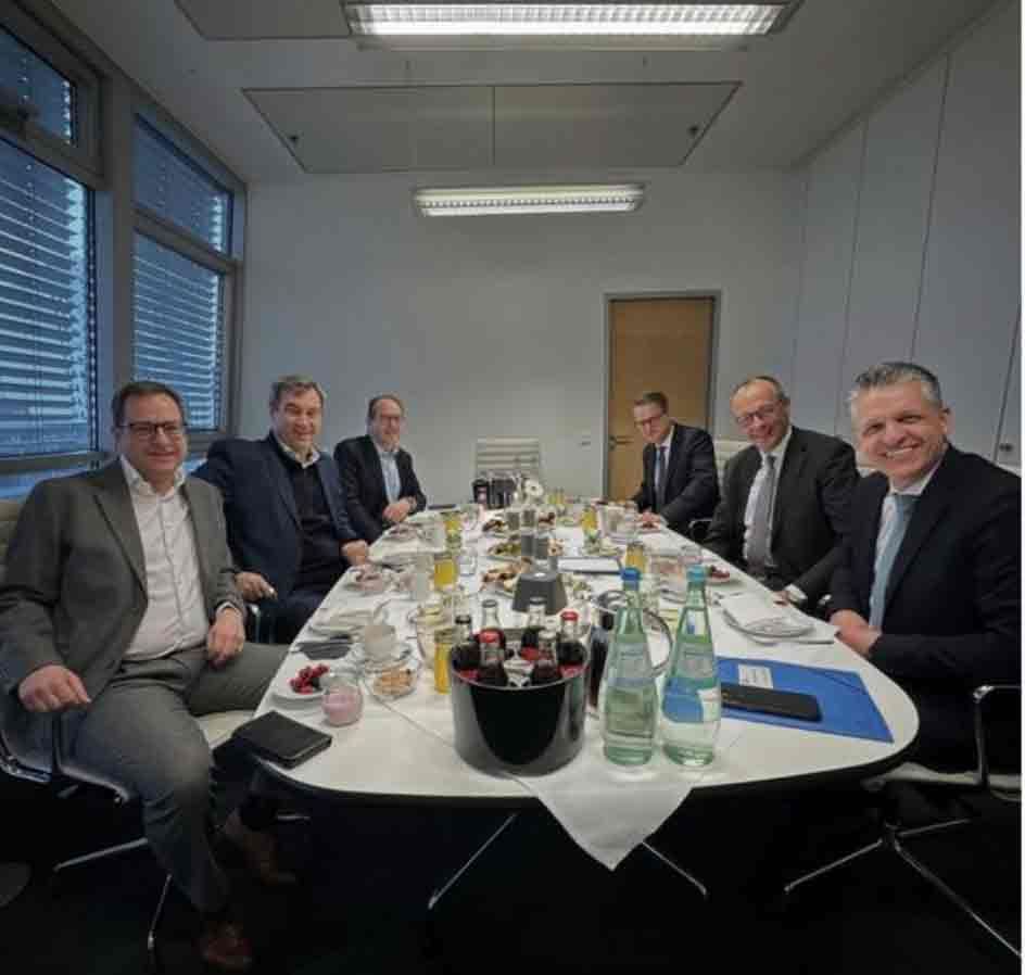 Mehrere männliche Personen sitzen in einem Besprechungsraum an einem Tisch mit Getränken und Unterlagen und schauen in die Kamera.