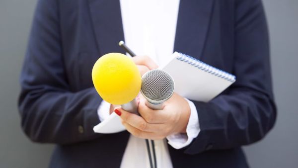 Person in dunklem Blazer hält zwei Mikrofone – eines mit gelbem Windschutz, eines silbern – sowie einen Notizblock und einen Stift in der Hand; Nahaufnahme vor neutralem Hintergrund.