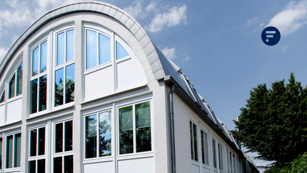 Halbrundes Gebäude mit Fensterfront, in dessen Fenstern sich Himmel, Woklen und Bäume spiegeln. Oben rechts ist das Logo fon Fink & Fuchs eingefügt.