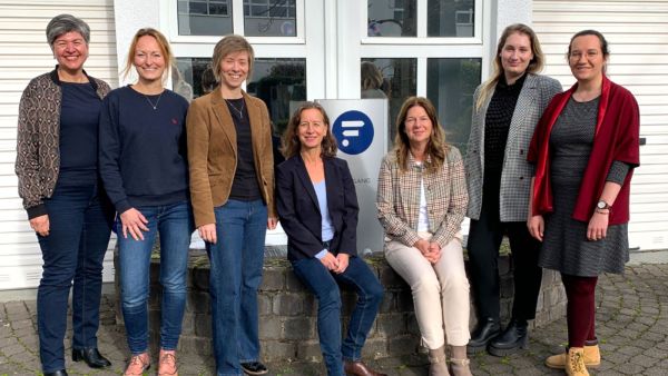 Sieben Frauen stehen und sitzen vor einem Gebäude mit großem Fenster und einem Schild mit blauem Logo. Sie lächeln in die Kamera und wirken wie ein Teamfoto in einer beruflichen oder organisatorischen Umgebung