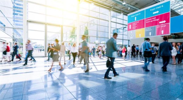 Die Eingangshalle eines internationalen Messe-Events: Menschen mit verschwommenen Konturenhasten durch ein sonnendurchflutetes Atrium, links oben sind Wegweiser zu den Ausstellungshallen zu sehen.