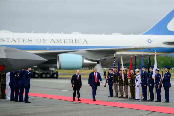Zwei Personen (links Wladimir Putin und rechts Donald Trump) gehen über einen roten Teppich auf einem Flughafen, flankiert von uniformierten Ehrenformationen, im Hintergrund steht ein großes Regierungsflugzeug.