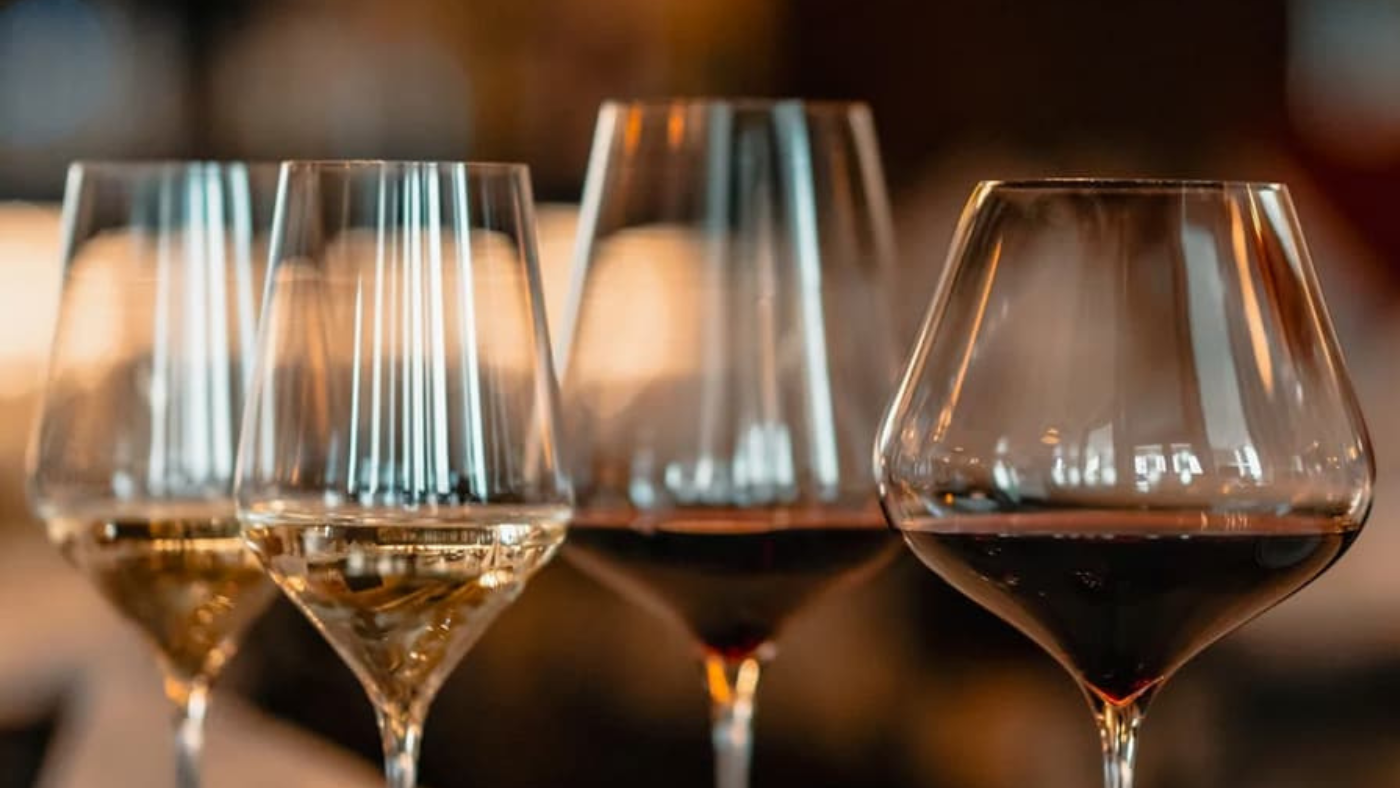 Nahaufnahme von vier Weingläsern auf einem Tisch, gefüllt mit Weiß- und Rotwein, vor einem warmen, unscharfen Hintergrund.